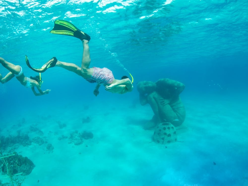 Two scuba divers swim beneath water towards a statue.