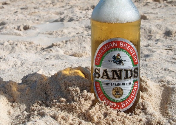 A bottle of Sands beer sticks out of the sand on a Bahamas beach. 