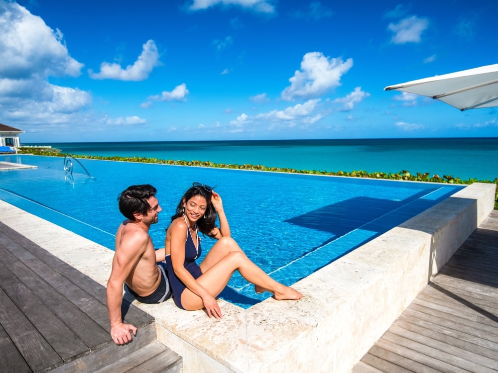 A couple sits on the edge of an infinity pool on a sunny day. 