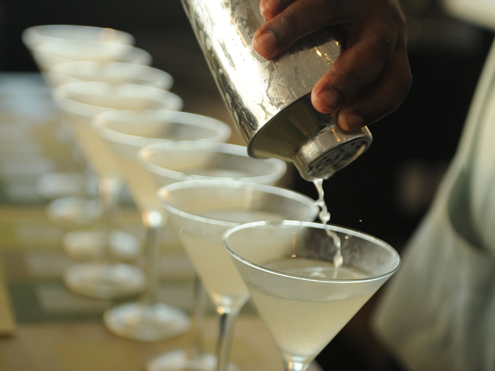 A freshly made cocktail is poured from a cocktail shaker into a row of martini glasses. 